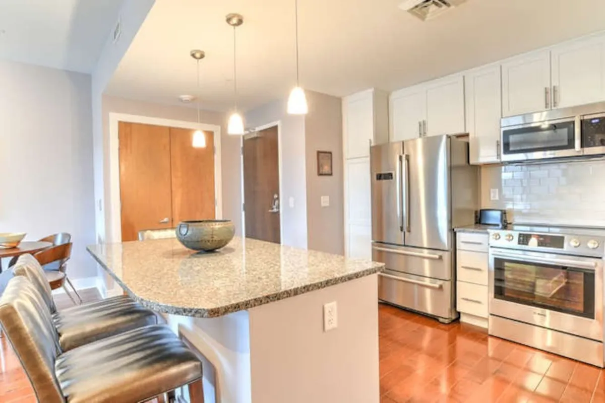 A bright, open-concept kitchen featuring a spacious granite countertop, sleek stainless steel appliances, pendant lighting, and comfortable bar stools. The space is set up for cooking, socializing, and casual dining, creating a welcoming hub for families or groups to gather after a day of city exploration.