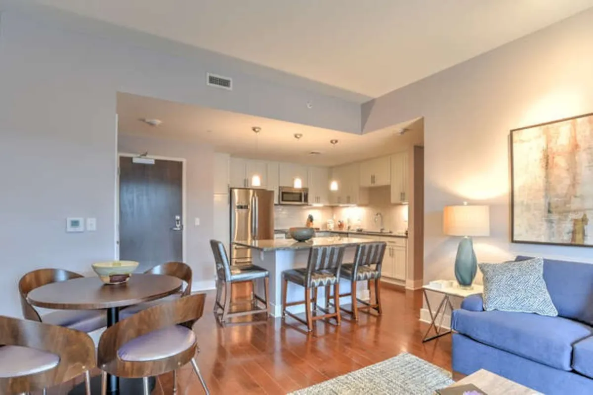 This wide-angle interior shot captures the heart of the open-concept living area, featuring a flowing layout connecting the kitchen island, dining table, and plush couch. Sunlight pours in through large east-facing windows, illuminating artwork and creating an inviting space ideal for socializing, dining, or relaxing. The image highlights multiple gathering zones and the seamless integration of living, dining, and cooking spaces.
