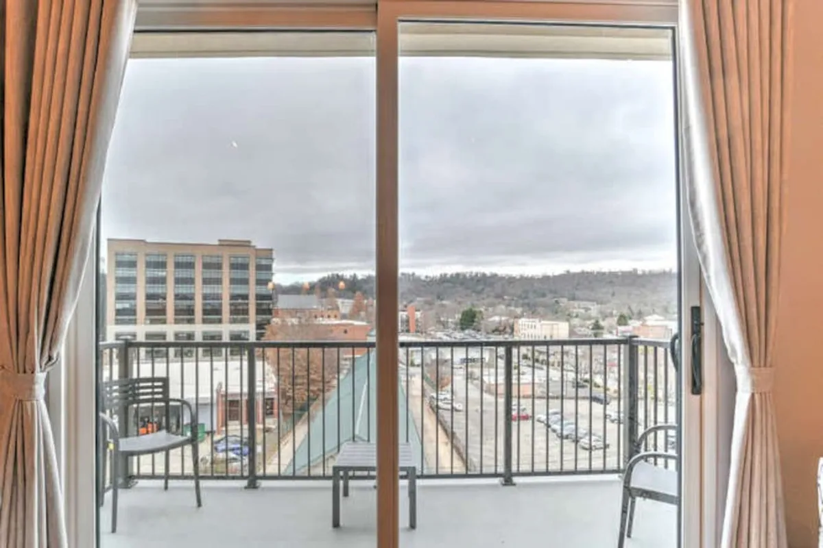 This exterior shot captures the apartment's balcony, featuring cozy outdoor seating and sheer curtains that frame a sweeping view of Asheville’s vibrant downtown. Early sunlight softly illuminates the scene, suggesting a perfect spot for morning coffee or evening relaxation while staying connected to urban energy.