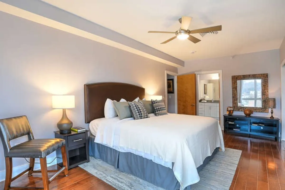 A serene interior view of a bedroom with a plush bed, a nightstand with a lamp for softly lit evenings, a small seating area, and a dressing table by the window. The morning sun illuminates the room, highlighting its calm and inviting vibe.