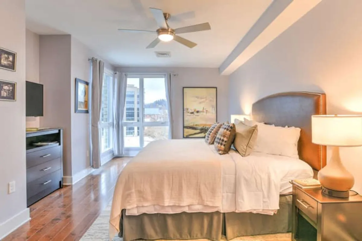 An inviting bedroom interior focused on a plush bed positioned near a large window. Soft natural light pours in, and the window hints at the city just beyond, suggesting the balcony’s proximity. Artful décor and a bedside lamp add to the serene, restful ambiance, evoking the transition from indoor comfort to outdoor enjoyment.
