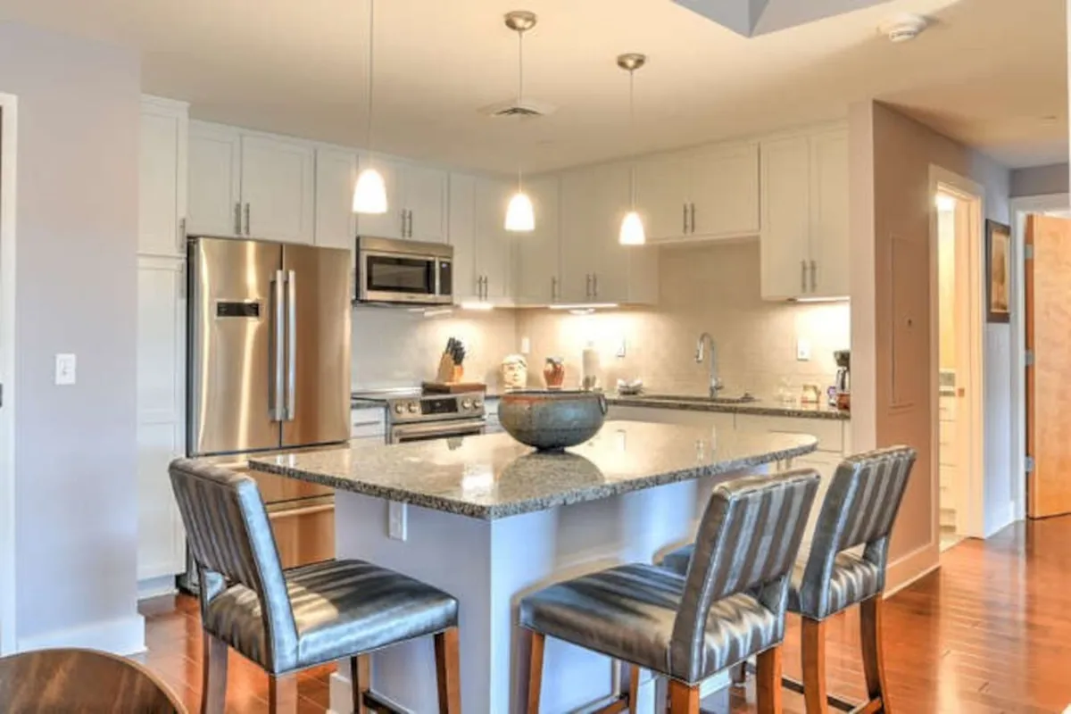 A stylish view of the modern kitchen area, focusing on the granite countertop, premium stainless steel appliances, pendant lighting, and bar stools arranged around the island. The scene conveys a welcoming atmosphere for cooking, entertaining, and casual dining—emphasizing the kitchen as a hub for connection and comfort.
