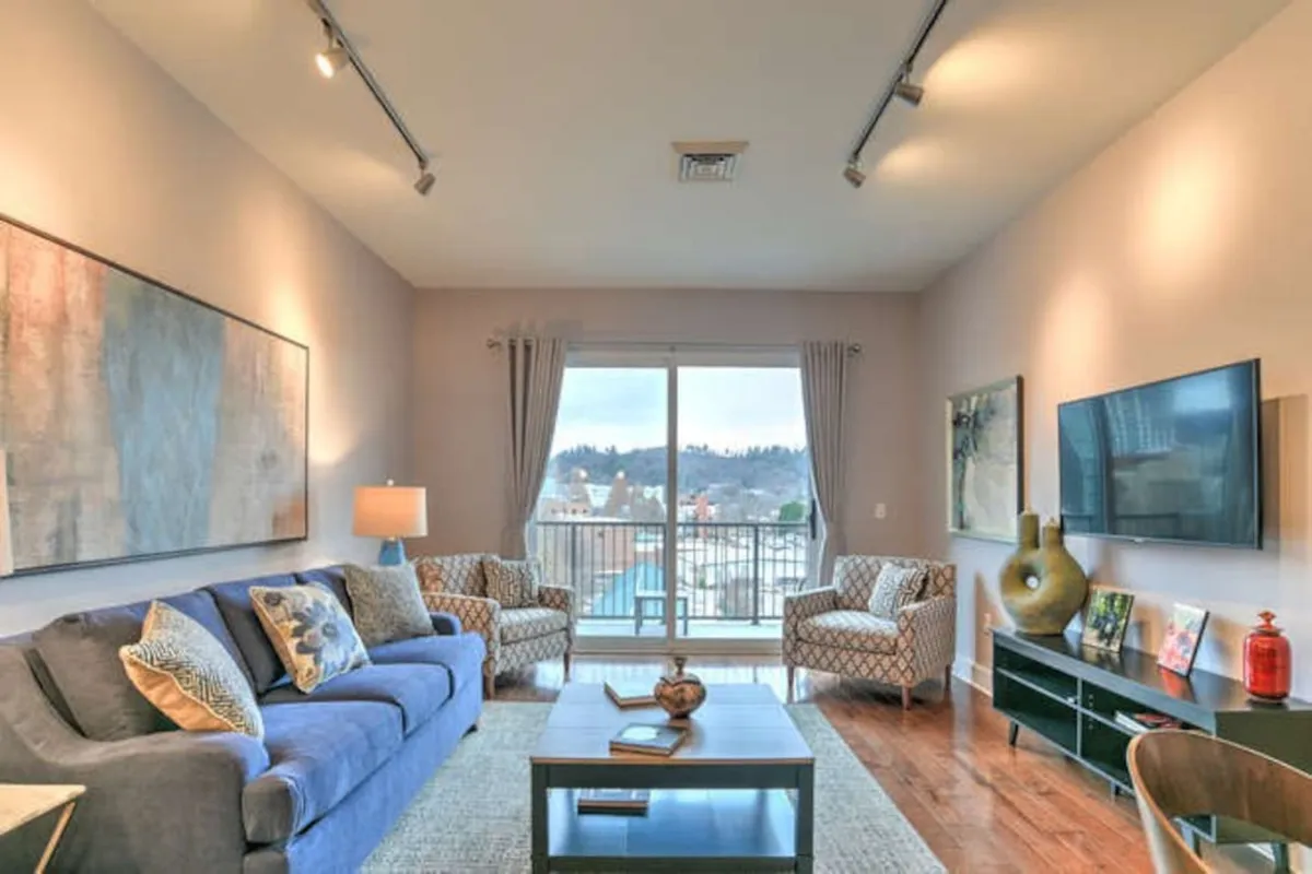The image showcases a stylish, comfortable living room with a plush sofa and coffee table, bathed in soft evening light from a large window. The window offers a scenic view—perfect for sunset watching—while curated artwork adorns the walls, and a flat-screen TV is ready for entertainment. The ambiance is warm and inviting, ideal for relaxing after a day in downtown Asheville.
